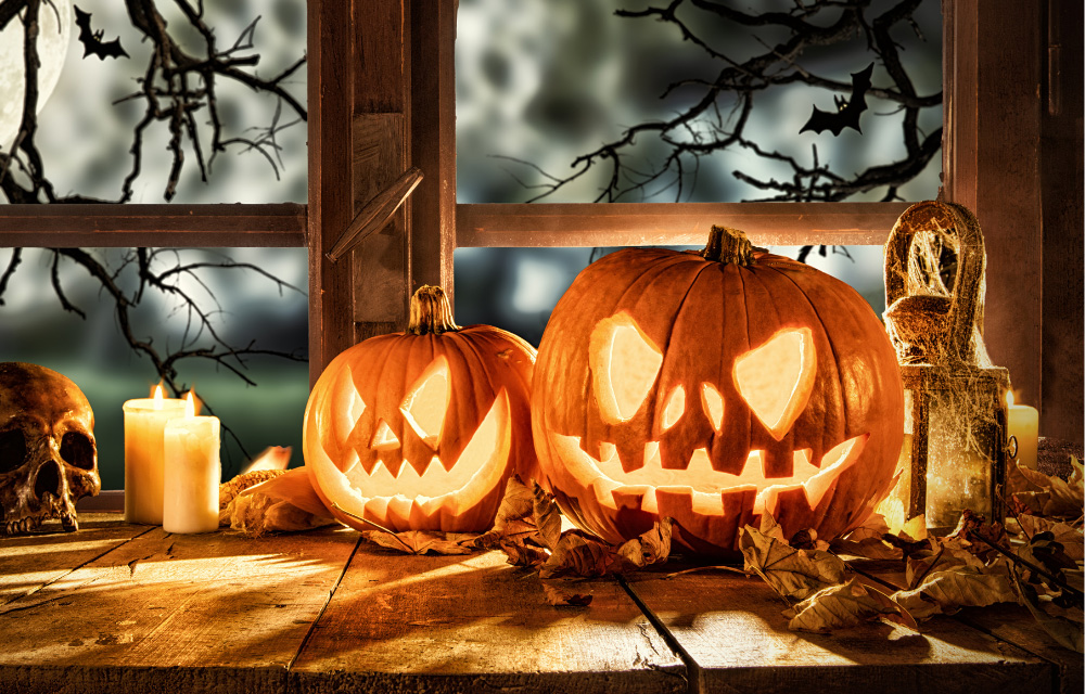 Schaurig-schöne Halloween-Deko: Zwei geschnitzte, leuchtende Kürbisse, Kerzen und ein Totenkopf auf einem rustikalen Holztisch vor dem Fenster.
