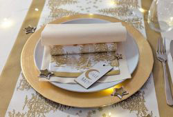 Ein einzelnes, festlich gedecktes Gedeck in Gold und Weiß. Auf dem goldenen Platzteller liegen Servietten, eine davon mit goldenem Waldmotiv. Dazu kleine goldene Sterne als Dekoration.
