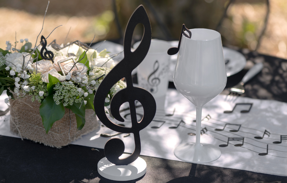 Tischdeko mit Musik-Motto: Großer schwarzer Notenschlüssel, weißes Weinglas und Blumen auf einem Noten-Tischläufer.