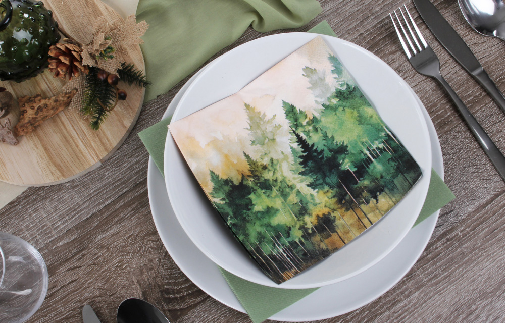 Naturnahe Gedeck-Idee: Eine Papierserviette mit Waldmotiv und eine rustikale Holzscheibe mit Tannenzapfen auf einem dunklen Holztisch.